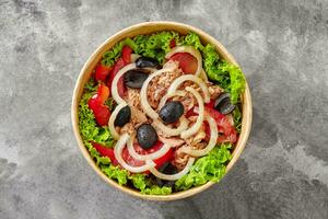 Salad with lettuce, tomatoes, bell pepper, canned tuna, marinated onion and black olives in cardboard bowl on gray stone surface photo