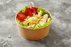 Poke bowl with lettuce, tomatoes, cucumbers, chicken fillet, corn and grated parmesan on gray stone surface photo