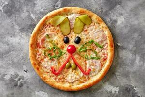 Funny pizza with mozzarella, ham and bunny muzzle of olives, cherry tomato, bell pepper, arugula and pickled cucumber photo