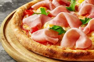 Closeup of pizza with melted mozzarella, ham, quail eggs, tomatoes and fresh greens on wooden board photo