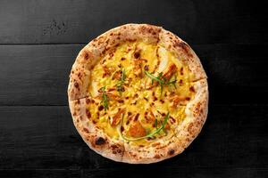 Pizza on thin dough with melted mozzarella, cheddar, chicken fillet and corn on black wooden background photo