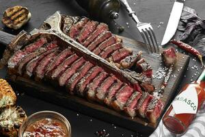 Sliced Steak T-bone lying on wooden board. Grilled vegetables in a pan grill. Black table with gray cloth photo