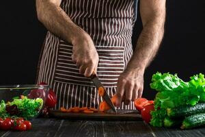 Young home cook man in apron slicing carrot with kitchen knife. photo