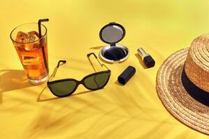 Straw beach woman's hat, beverage with ice in a glass and sun glasses on yellow background with shadow from a palm leaf. photo