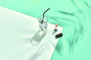 High angle view of lipstick and drinking glass with ice on table. Blue and white background with shadow from a palm leaf photo