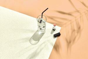 High angle view of lipstick and drinking glass with ice on table. Beige and white background with shadow from a palm leaf photo