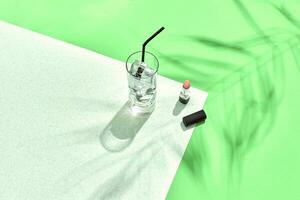 alto ángulo ver de lápiz labial y Bebiendo vaso con hielo en mesa. verde y blanco antecedentes con sombra desde un palma hoja foto