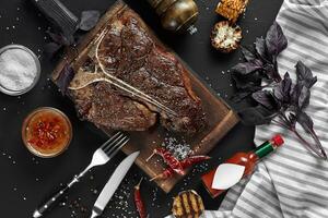 Succulent grilled large t-bone steak garnished with herbs, corn and salt with fork and knife beside it on cutting board photo