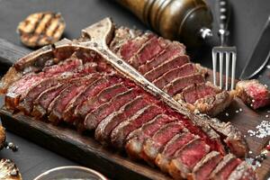 Sliced Steak T-bone lying on wooden board. Grilled vegetables in a pan grill. Black background. Close-up photo