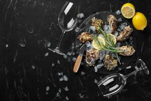 Fresh closed oysters, ice, lemon on a round slate and two empty glasses are on a black stone textured background. Top view with copy space. Close-up. photo