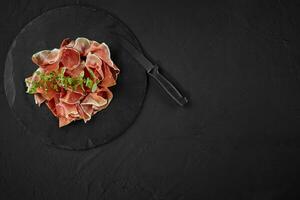 Sliced jamon with herbs and a knife are on black stone slate board against a dark grey background. Close-up shot. Top view. Copy space. photo