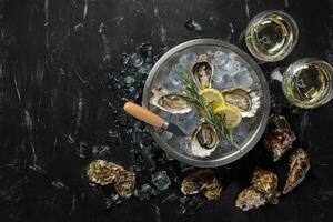 Fresh opened oysters, ice, lemon on a round metal plate and champagne are on a black stone textured background. Top view with copy space. Close-up. photo