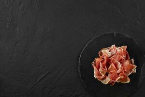 Gourmet, manually sliced jamon on black stone slate board against a dark grey background. Close-up shot. Top view. Copy space. photo