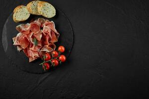 Sliced jamon, cherry tomatoes, baguette, herbs on black stone slate board against a dark grey background. Close-up shot. Top view. Copy space. photo