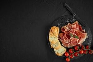 Sliced jamon, cherry tomatoes, baguette, herbs, spices and knife on black stone slate board against a dark grey background. Close-up shot. Top view. photo