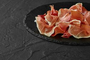 Gourmet, manually sliced jamon on black stone slate board against a dark grey background. Close-up shot. Side view. Copy space. photo