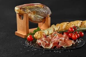 Jamon carved from pig leg on a jamonera stand, cherry tomatoes, baguette, herbs, spices on black slate board. Dark background. Close-up. Side view. photo