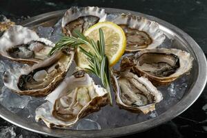 Fresh opened oysters, ice and lemon on a round metal plate, black stone textured background. Side view with copy space. Close-up shot. photo