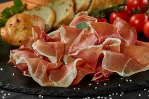 Sliced jamon, cherry tomatoes, baguette, herbs and spices on black stone slate board against a dark grey background. Close-up shot. Side view. photo