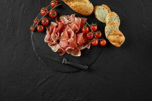 Sliced jamon, cherry tomatoes, sliced baguette and a knife on black stone slate board against a dark grey background. Close-up. Top view. Copy space. photo