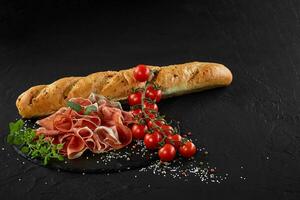 Sliced jamon, cherry tomatoes, herbs and spices on black stone slate board. Baguette is laying nearby on dark grey background. Close-up. Side view. photo