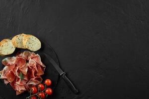 Sliced jamon, cherry tomatoes, baguette, herbs and knife on black stone slate board against dark grey background. Close-up. Top view. Copy space. photo