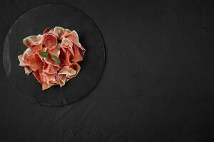 Gourmet, manually sliced jamon with herbs on black stone slate board against a dark grey background. Close-up shot. Top view. Copy space. photo