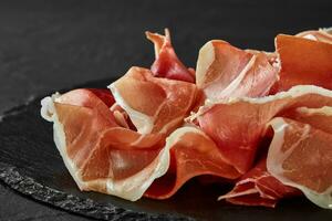 Gourmet, manually sliced jamon on black stone slate board against a dark grey background. Close-up shot. Side view. Copy space. photo