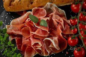 Sliced jamon, cherry tomatoes, baguette, herbs and spices on black stone slate board against a dark grey background. Close-up shot. Top view. photo
