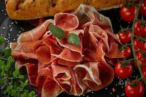 Sliced jamon, cherry tomatoes, baguette, herbs and spices on black stone slate board against a dark grey background. Close-up shot. Top view. photo