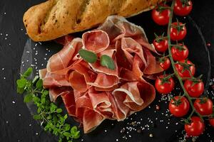 Sliced jamon, cherry tomatoes, baguette, herbs and spices on black stone slate board against a dark grey background. Close-up shot. Top view. photo