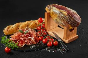 Jamon carved from pig leg on jamonera stand, cherry tomatoes, herbs on black stone slate board, baguette and knife on dark background. Close-up. photo