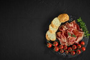 Sliced jamon, cherry tomatoes, baguette, herbs and spices on black stone slate board against a dark grey background. Close-up shot. Top view. photo