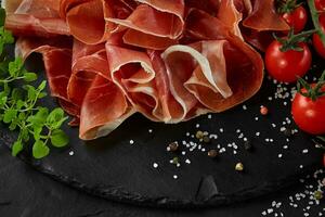 Sliced jamon, cherry tomatoes, herbs and spices on black stone slate board against a dark grey background. Close-up shot. Top view. photo