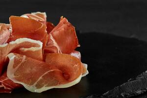 Gourmet, manually sliced jamon on black stone slate board against a dark grey background. Close-up shot. Side view. Copy space. photo