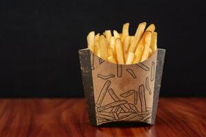 French fries in paper box on wooden table, black background, selective focus photo