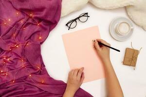 Top view of girl's hand writing in notepad placed on white surface with coffee cup and other items. Mock up photo