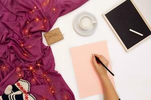 Top view of girl's hand writing in notepad placed on white surface with coffee cup and other items. Mock up photo