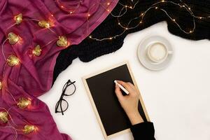 Top view of girl's hand writing on small black wooden board on white surface with coffee cup and other items. Mock up photo