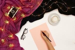 Top view of girl's hand writing in notepad placed on white surface with coffee cup and other items. Mock up photo