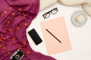 A cup of coffee, black smart, pink paper with new years goals. Scarf and Christmas lights on white background. photo