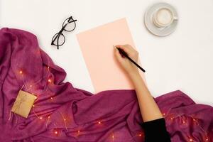 Top view of girl's hand writing in notepad placed on white surface with coffee cup and other items. Mock up photo