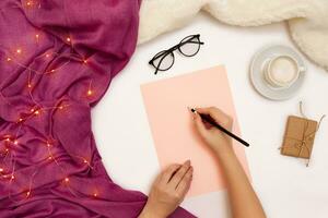 Top view of girl's hand writing in notepad placed on white surface with coffee cup and other items. Mock up photo