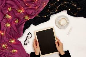 Top view of girl's hand writing on small black wooden board on white surface with coffee cup and other items. Mock up photo