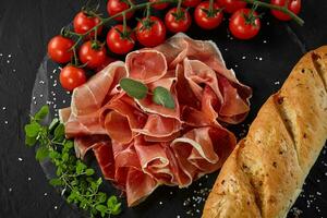 Sliced jamon, cherry tomatoes, baguette, herbs and spices on black stone slate board against a dark grey background. Close-up shot. Top view. photo