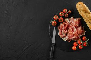 Sliced jamon, cherry tomatoes, baguette and a knife on black stone slate board against a dark grey background. Close-up shot. Top view. Copy space. photo