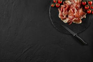 Sliced jamon, cherry tomatoes and a knife on black stone slate board against a dark grey background. Close-up shot. Top view. Copy space. photo