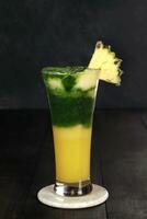 pineapple juice with vegetables and pineapple chunks garnish. has yellow and green gradations. served in a clear glass with a white and dark background photo