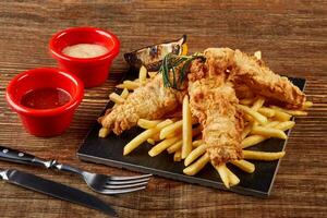 Delicious golden batter deep fried fish fillets and French fries, served on black board on wooden worktop, view from above. photo