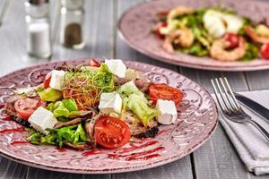 Healthy mixed Greek salad served on a pink plate with silver fork containing crisp leafy greens, microgreen, feta, onion, tomato and sliced beef photo